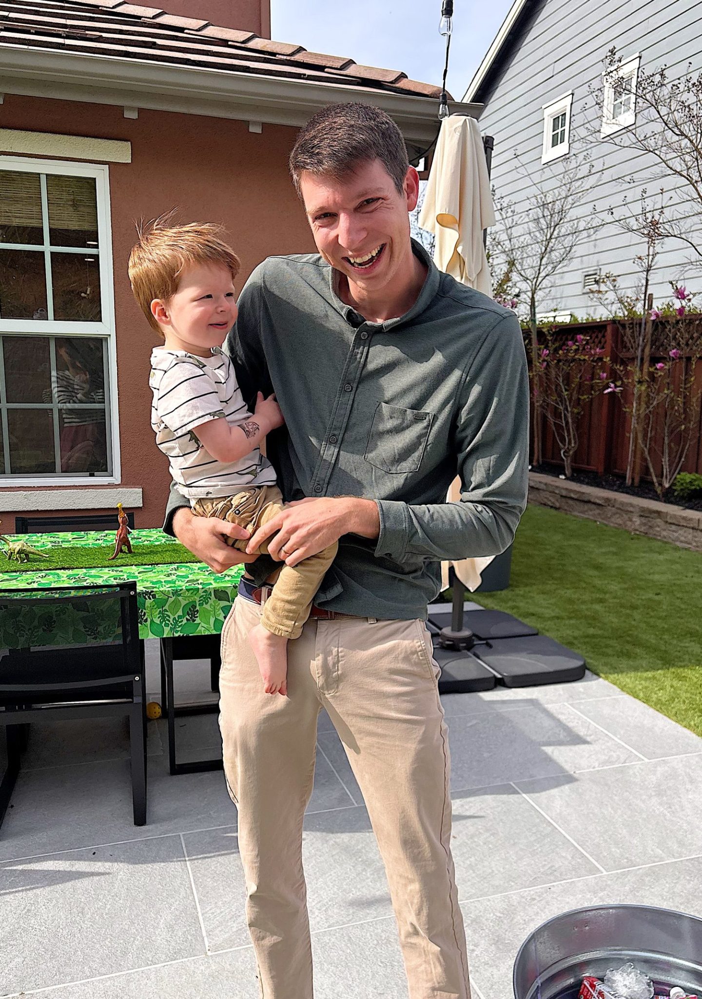 An adult man stands outdoors holding a young child in his arms, both smiling, with patio furniture and greenery in the background.