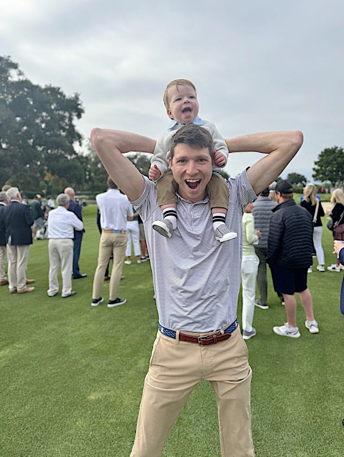 A man stands on a golf course with a baby sitting on his shoulders; the baby appears to be crying while people gather in the background.