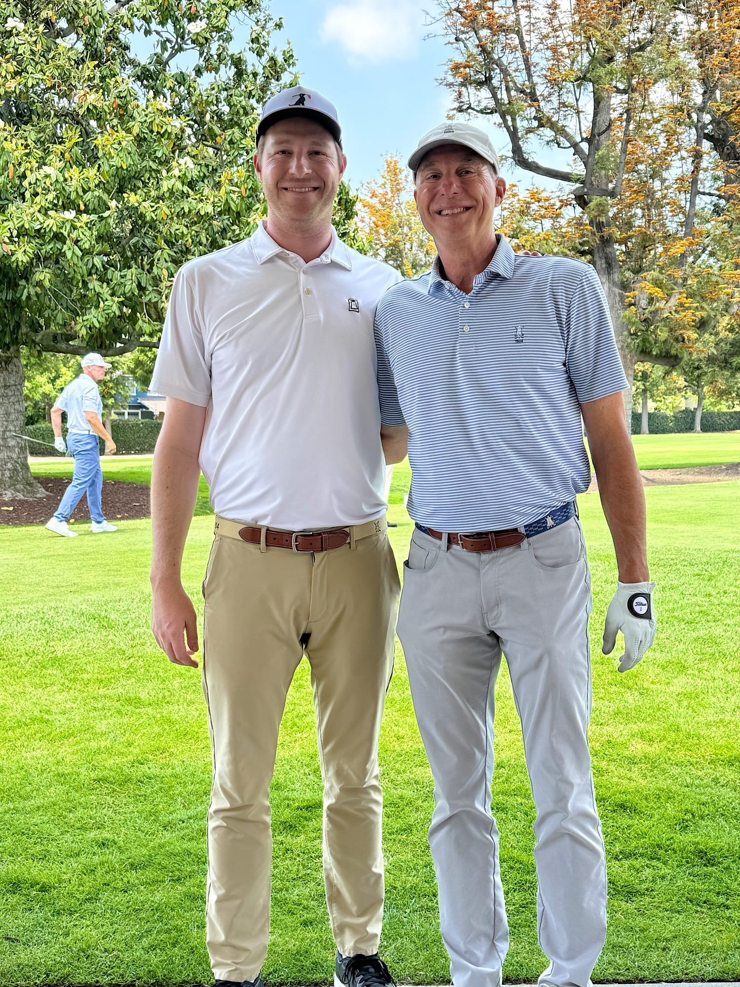 Two men in golf attire stand side by side on a golf course, smiling at the camera, with trees and another golfer in the background.