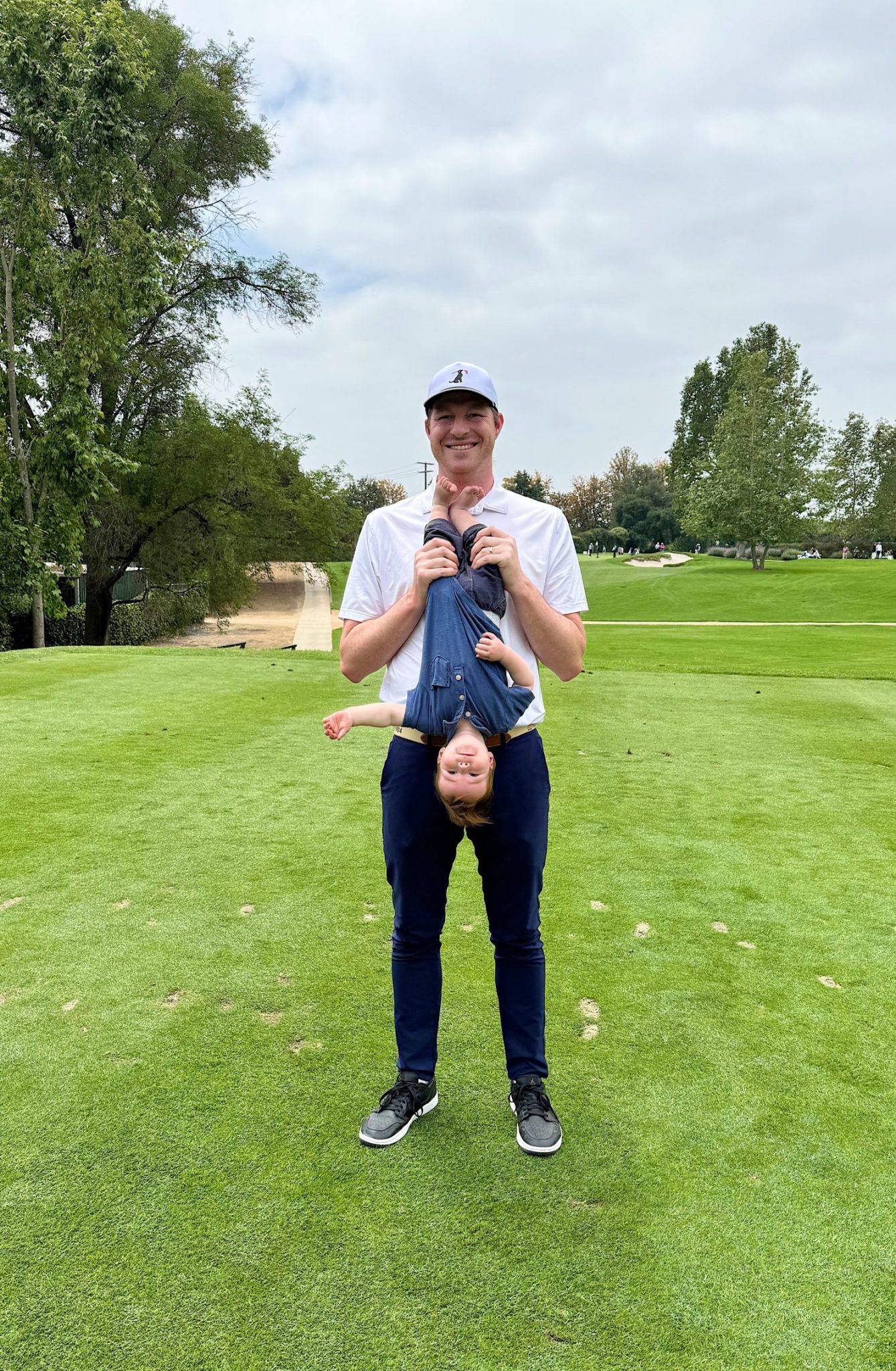 A man stands on a golf course holding a smiling child upside down by the ankles.