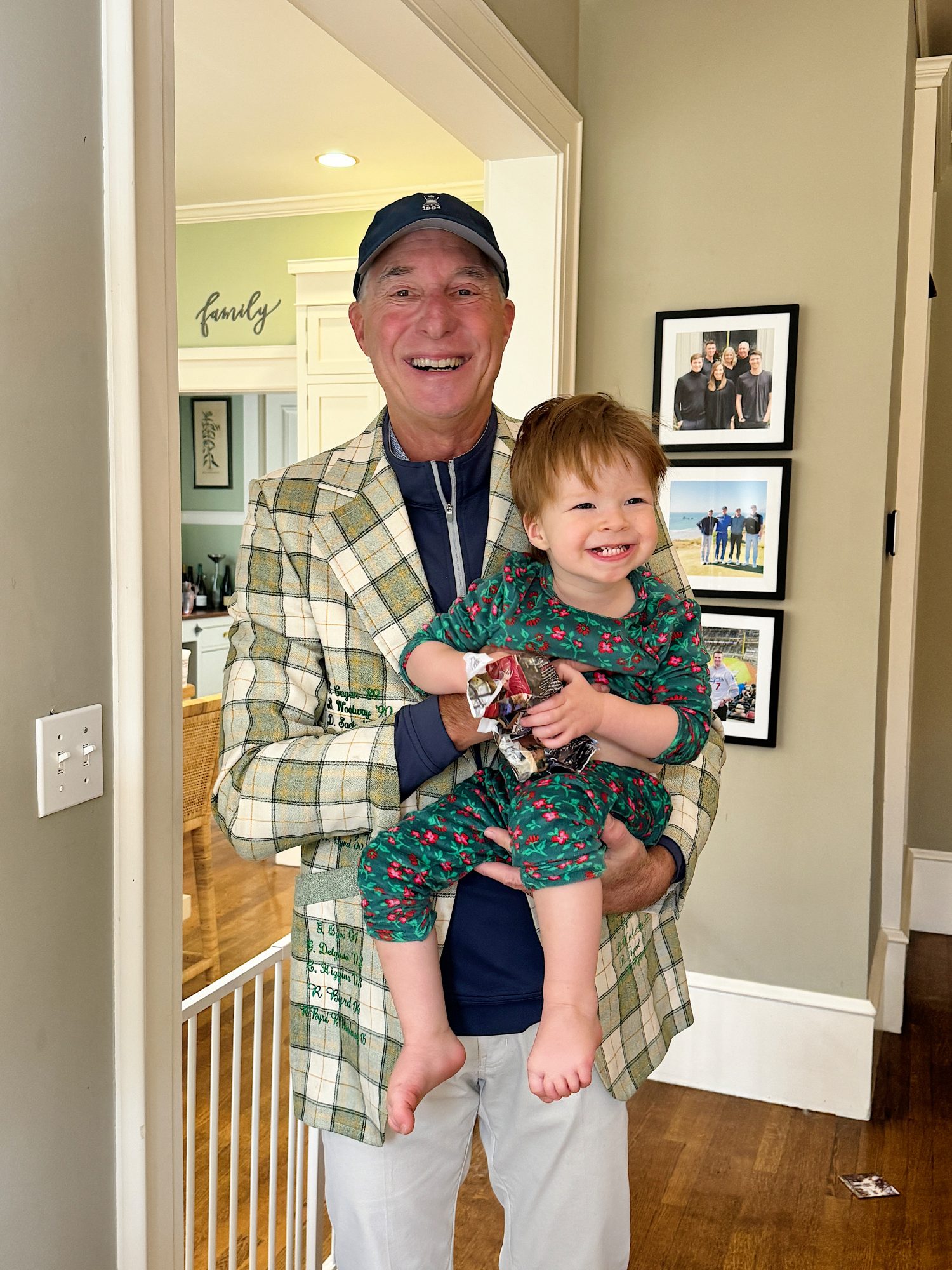 An older man in a plaid jacket and hat holds a smiling toddler in patterned pajamas, standing in a home near framed photos and a baby gate.