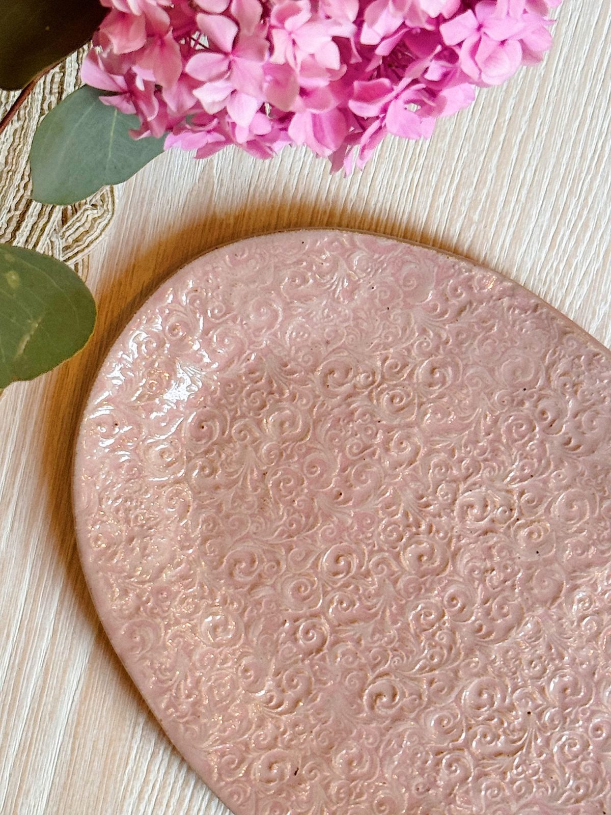 A pink, oval-shaped ceramic plate with an embossed swirl pattern sits on a light wood surface next to pink hydrangea flowers.