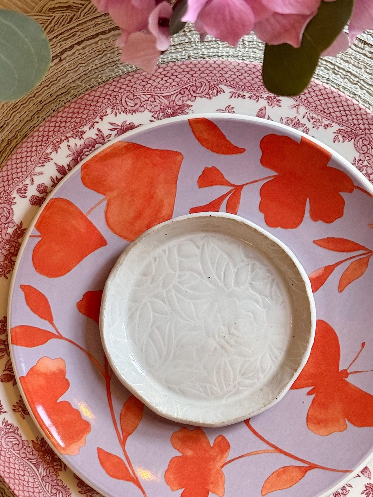 A small white textured dish rests on a larger plate with orange flower patterns, set on a patterned placemat with pink flowers visible at the top.