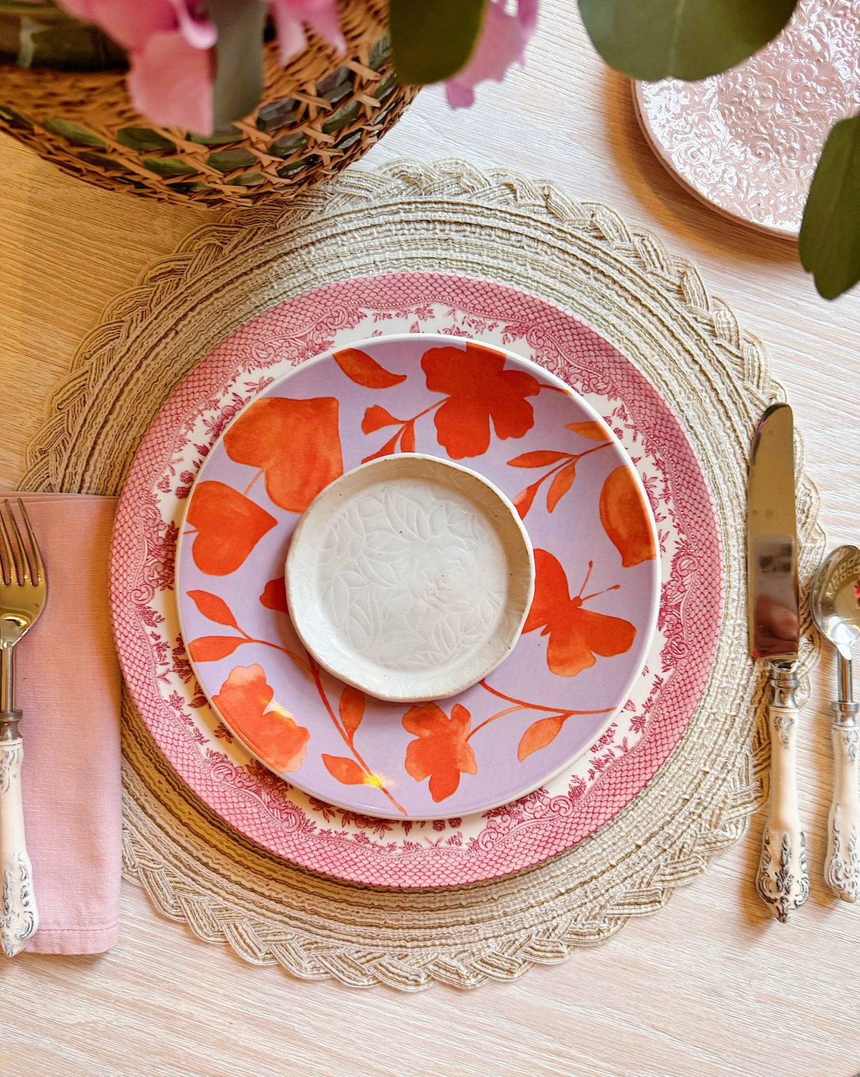 A place setting with a woven placemat, pink floral plate, red and pink floral plate, small white bowl, pink napkin, fork, knife, and spoon on a light wooden table.