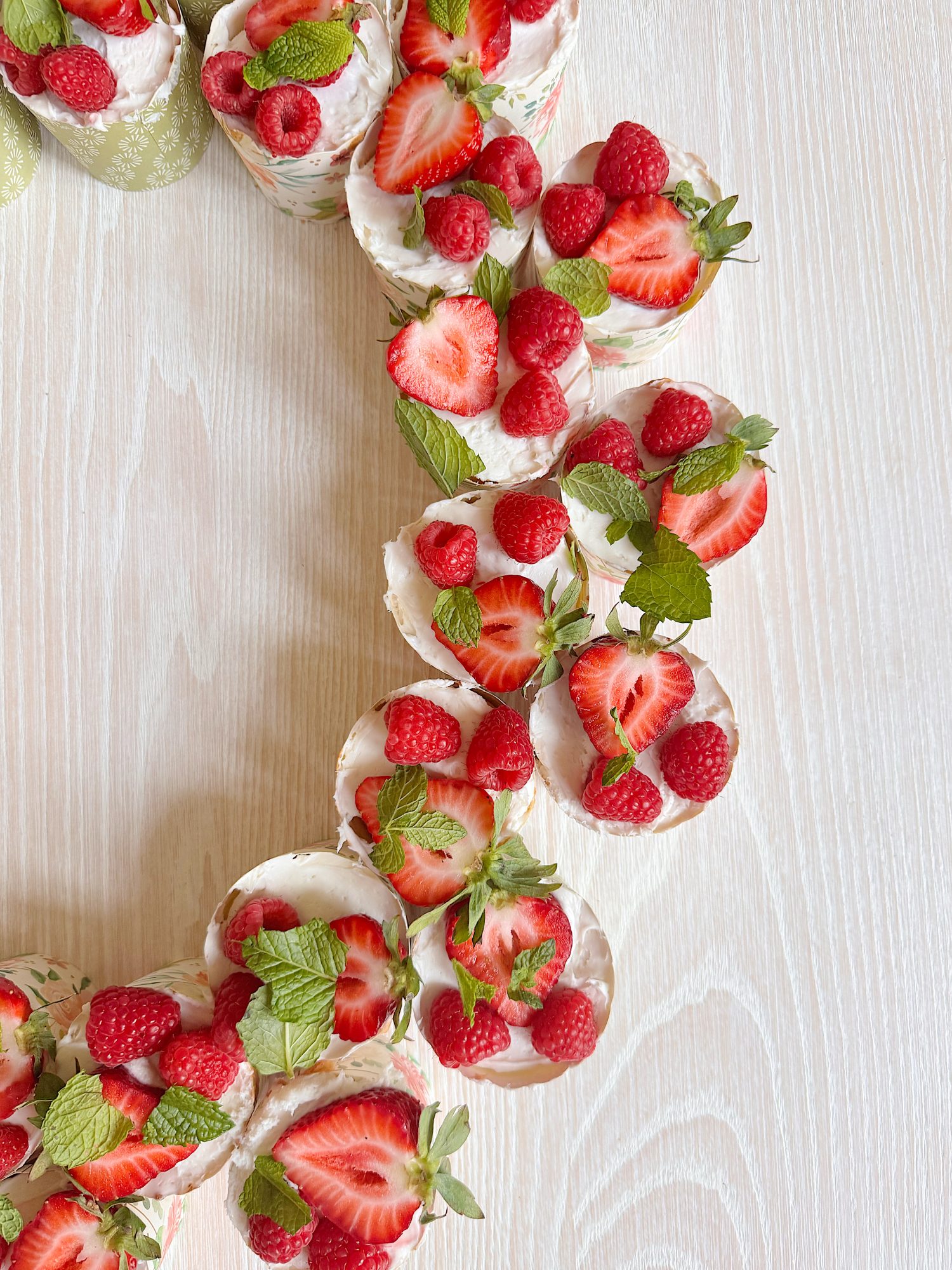 Cupcakes arranged in a curved line, topped with white frosting, fresh strawberries, raspberries, and mint leaves on a light wooden surface.