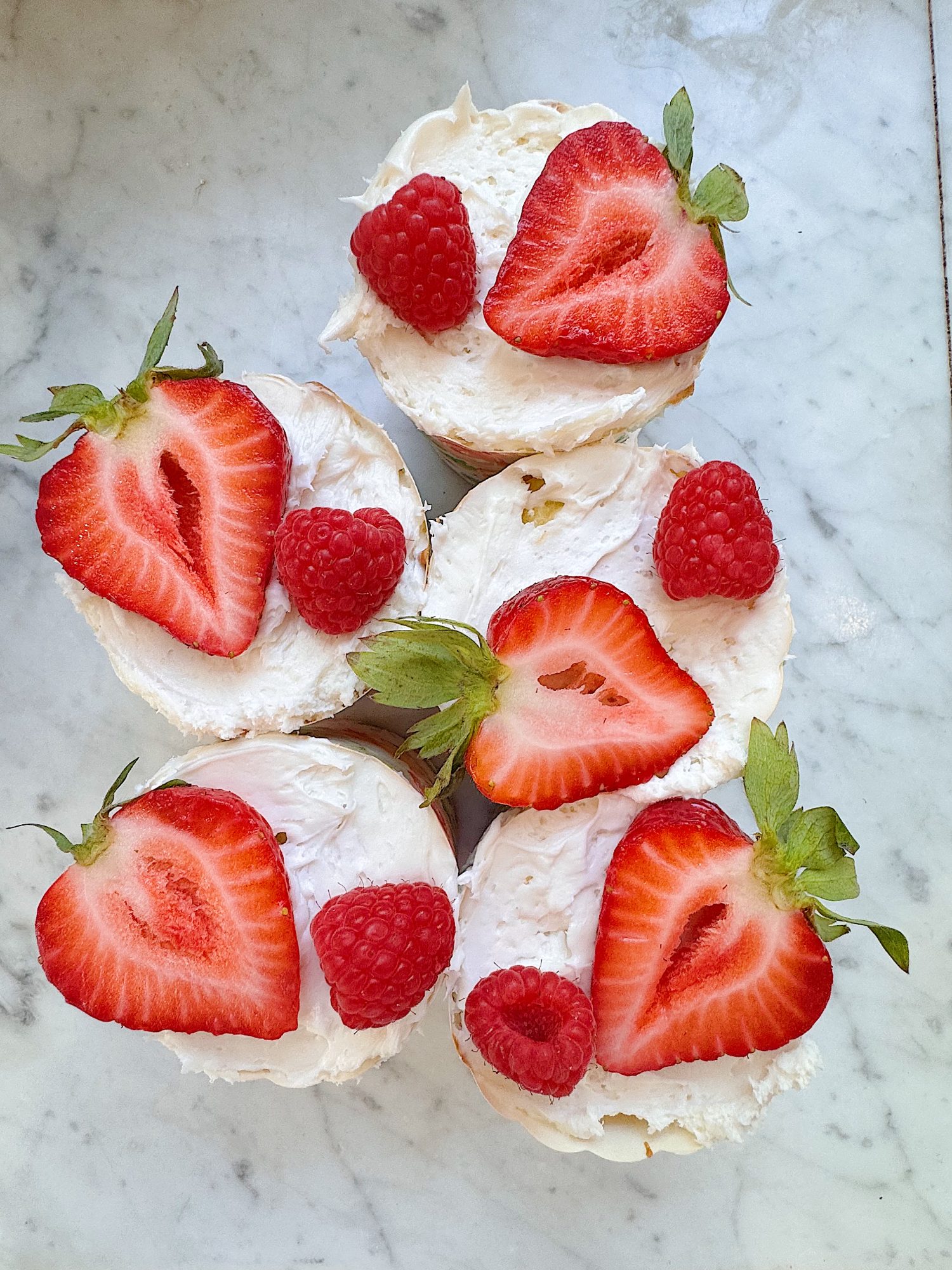 Four mini cakes topped with whipped cream, halved strawberries, and whole raspberries are arranged on a white marble surface.