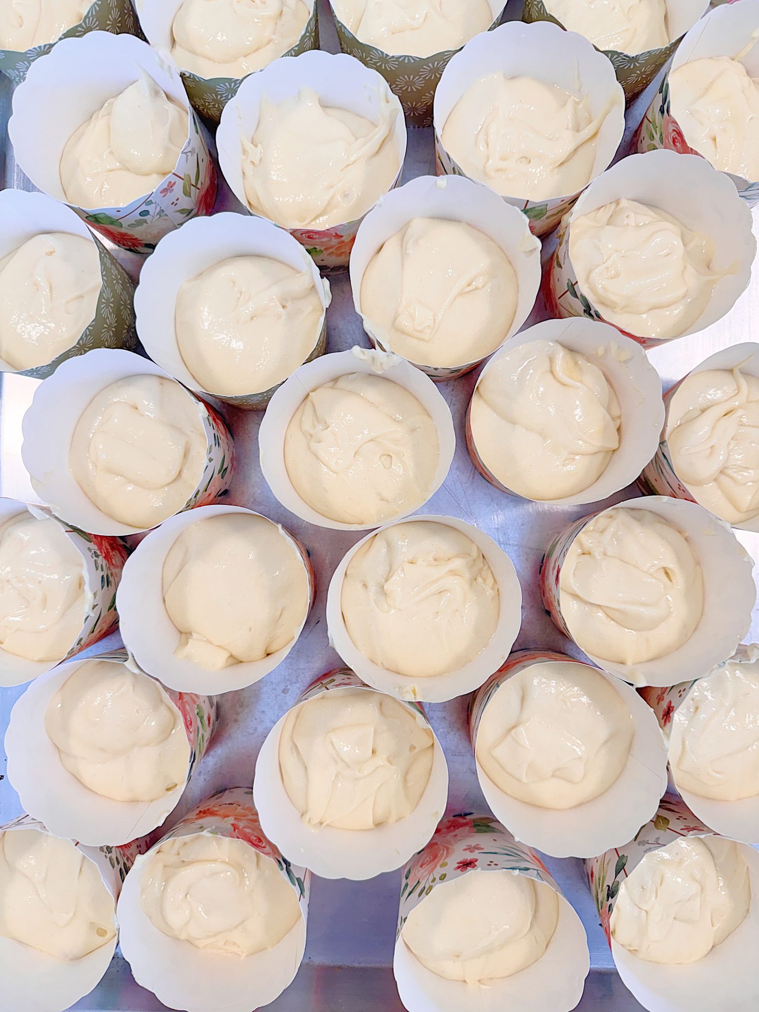 Rows of paper cupcake liners filled with pale, creamy batter arranged closely together on a flat surface.