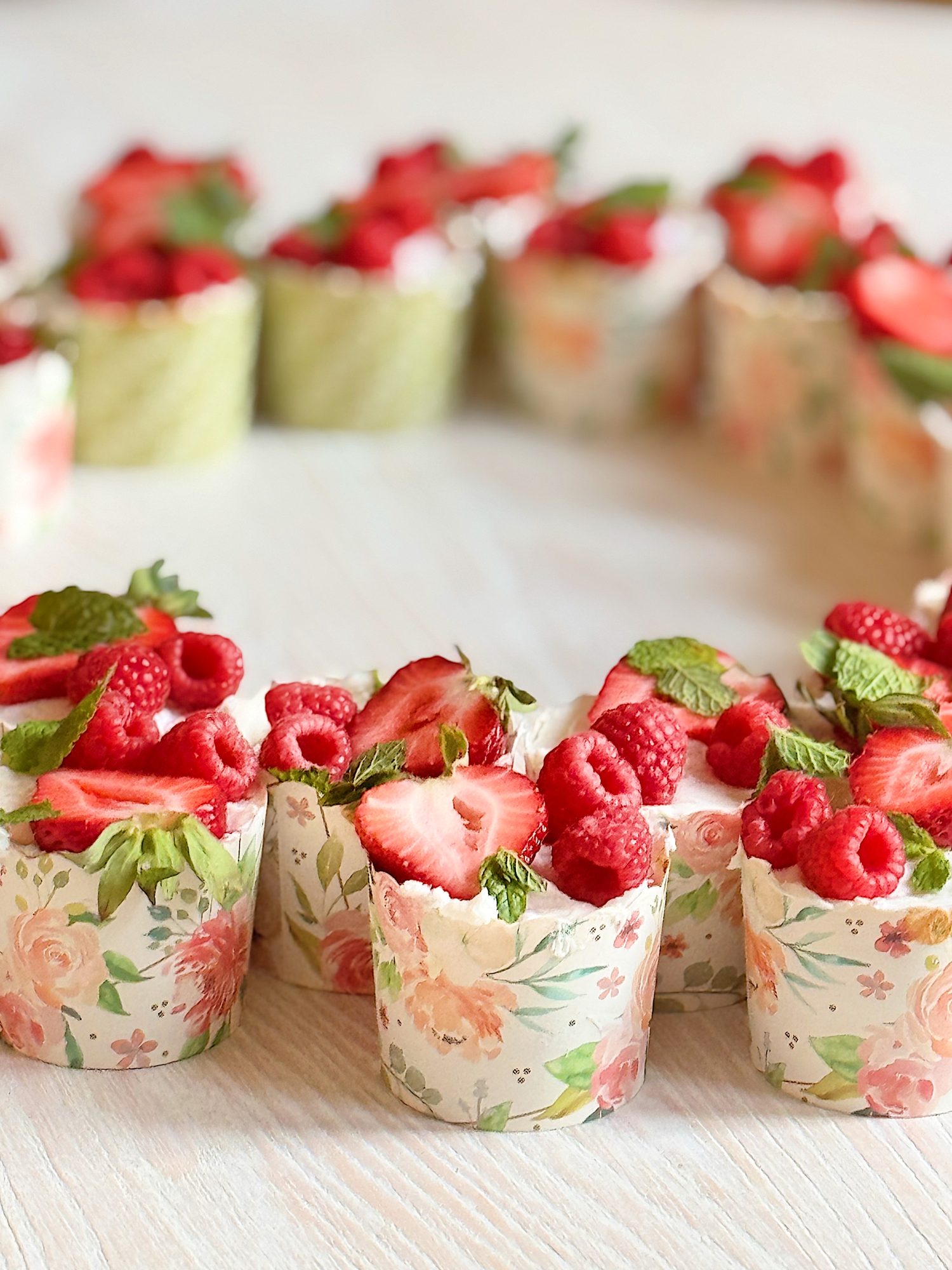 Small floral-patterned dessert cups filled with whipped cream, raspberries, sliced strawberries, and mint leaves, arranged in a ring shape on a light-colored surface.