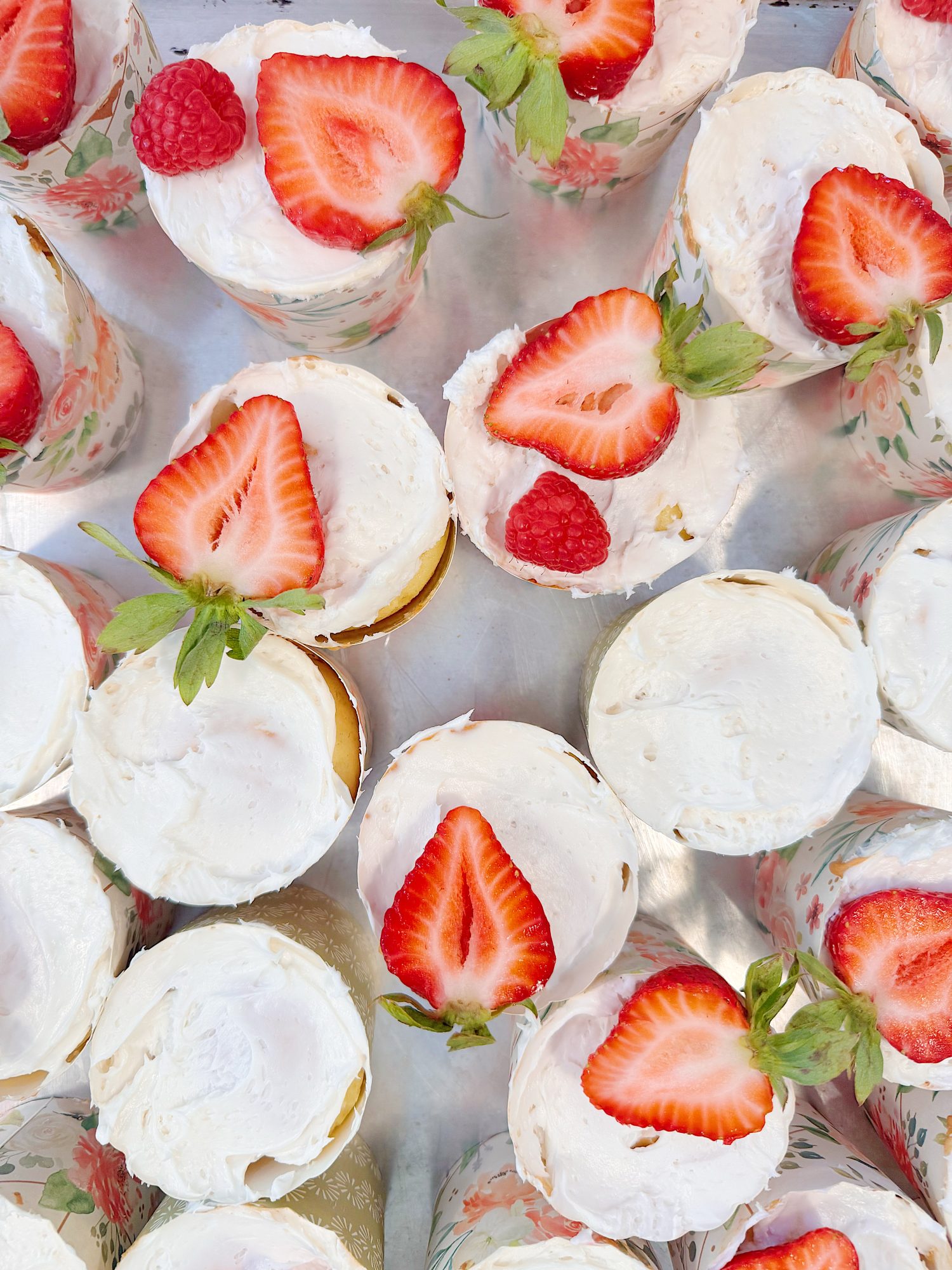 Cakes with white frosting are topped with halved strawberries and raspberries, arranged in floral-patterned wrappers on a tray.