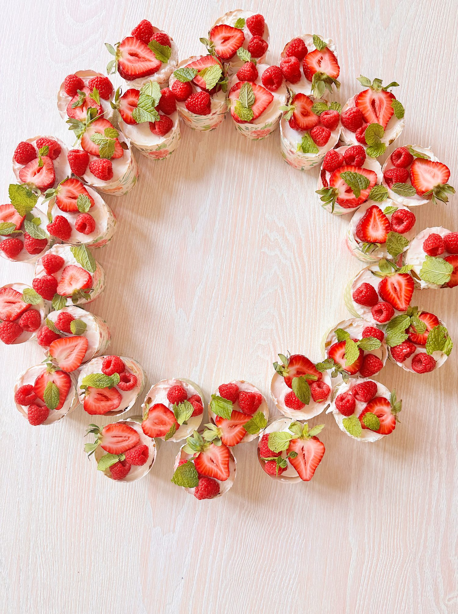A circular arrangement of cupcakes topped with strawberries, raspberries, and mint leaves on a light wooden surface.