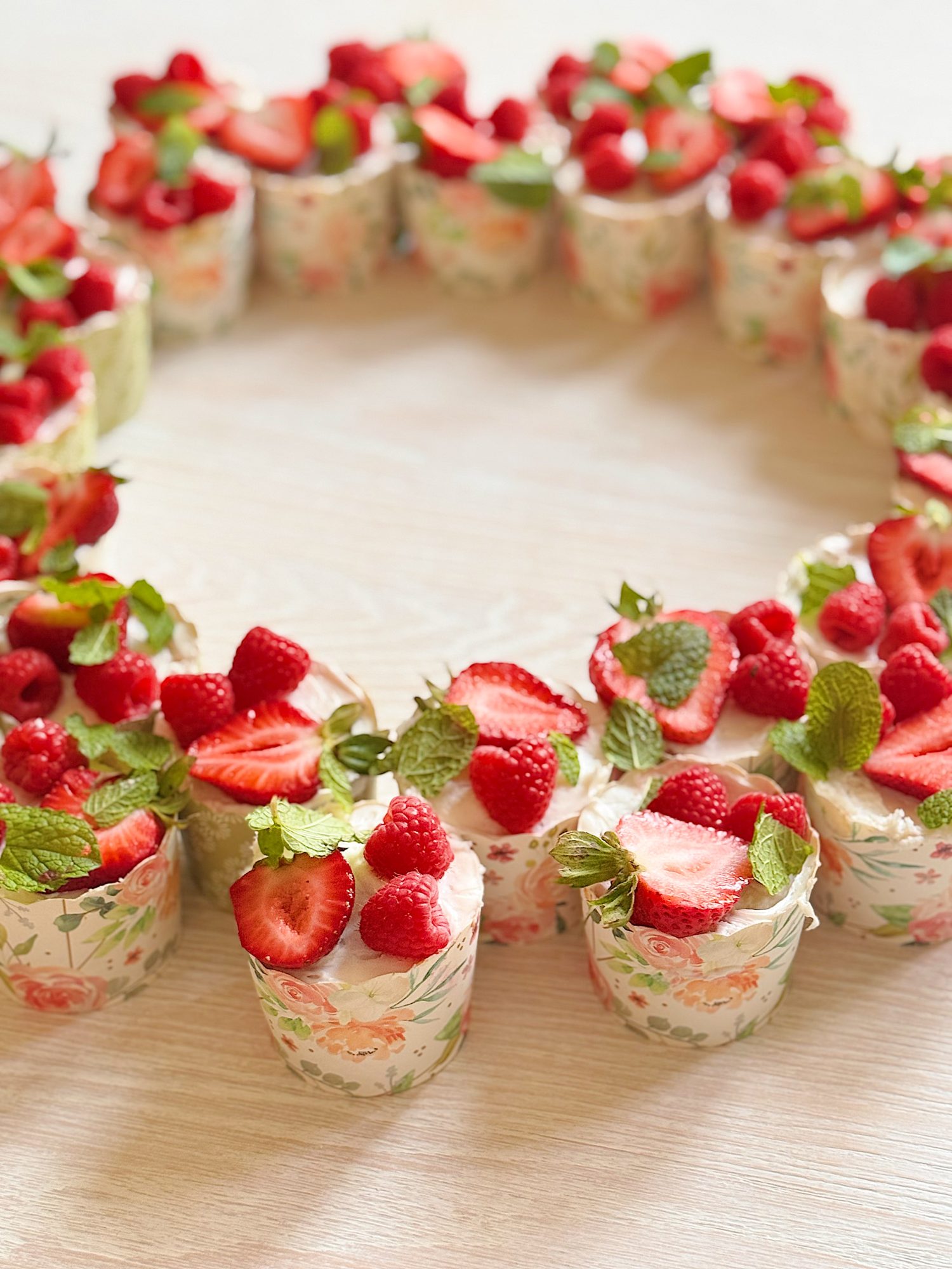 A group of small floral-patterned dessert cups topped with strawberries, raspberries, and mint leaves arranged in a circular pattern on a light wooden surface.
