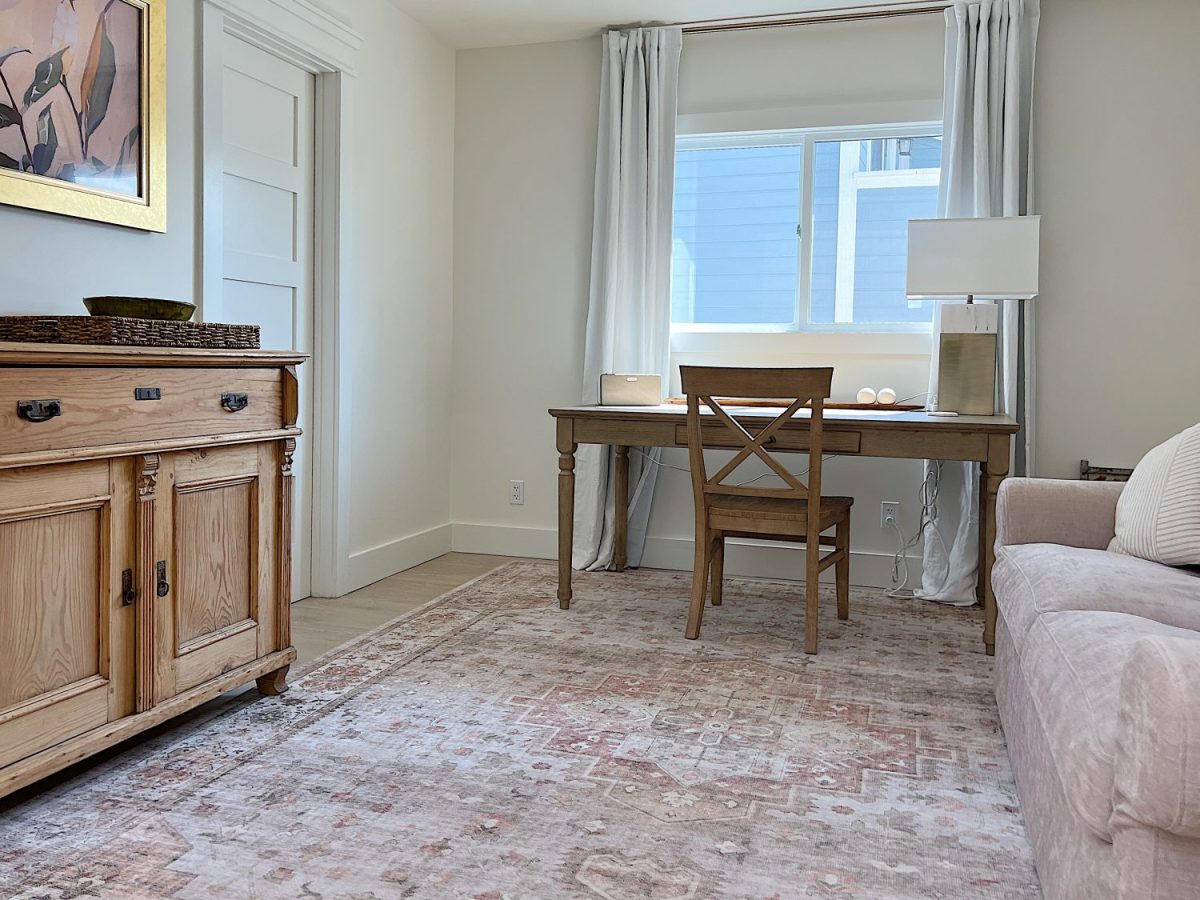 A home office with a wooden desk and chair, a lamp, a sideboard, a beige sofa, and a large window with sheer curtains letting in natural light.