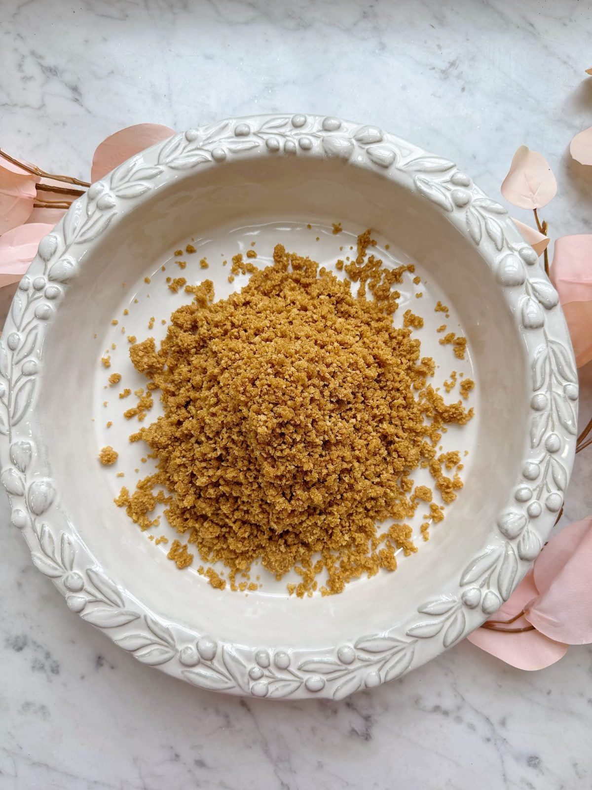 A white decorative bowl containing a mound of finely ground brown crumbs sits on a marble surface with pink leaves around the edges.
