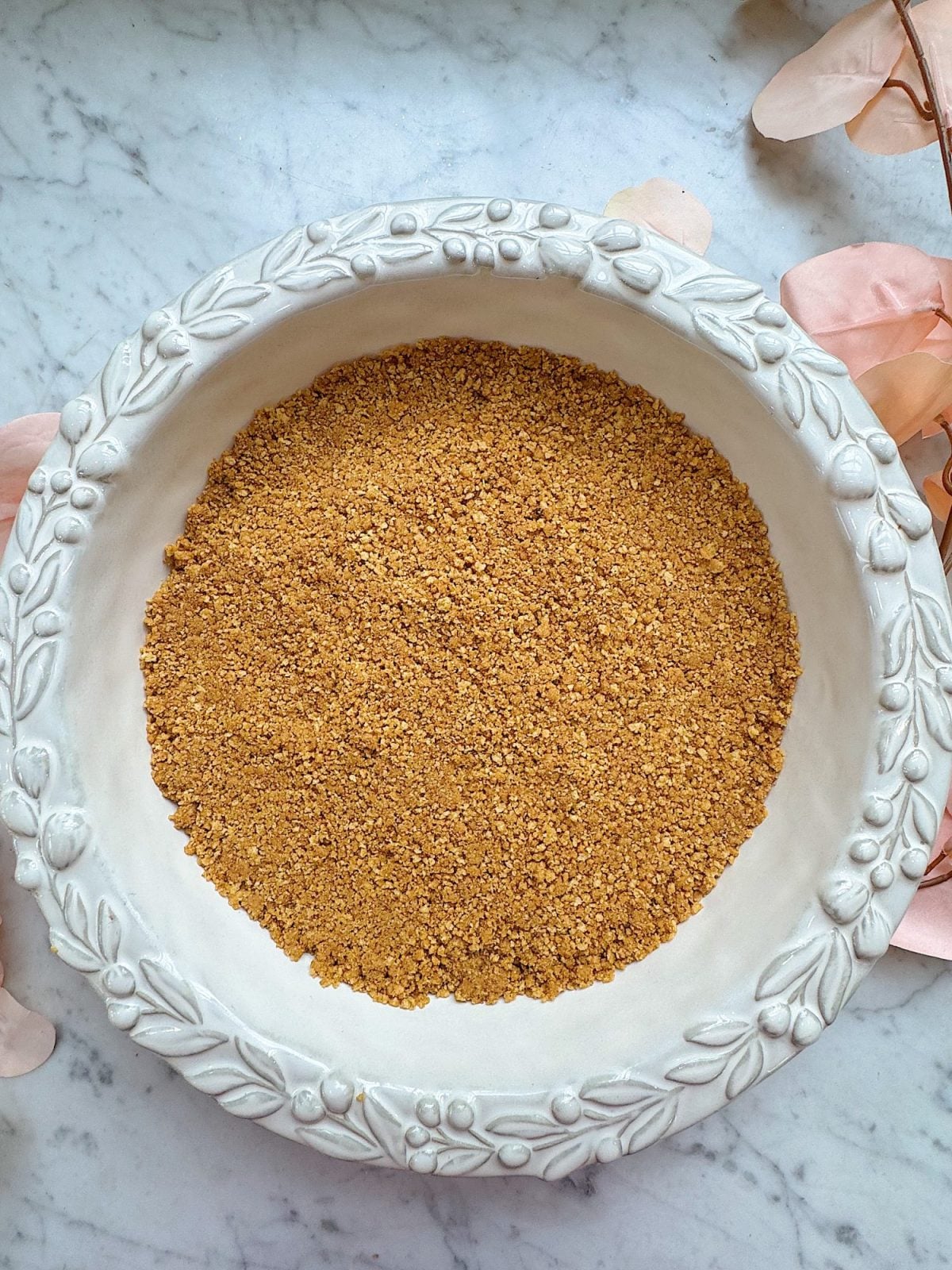 A white ceramic bowl filled with finely ground brown powder sits on a marble surface, surrounded by light pink leaves.