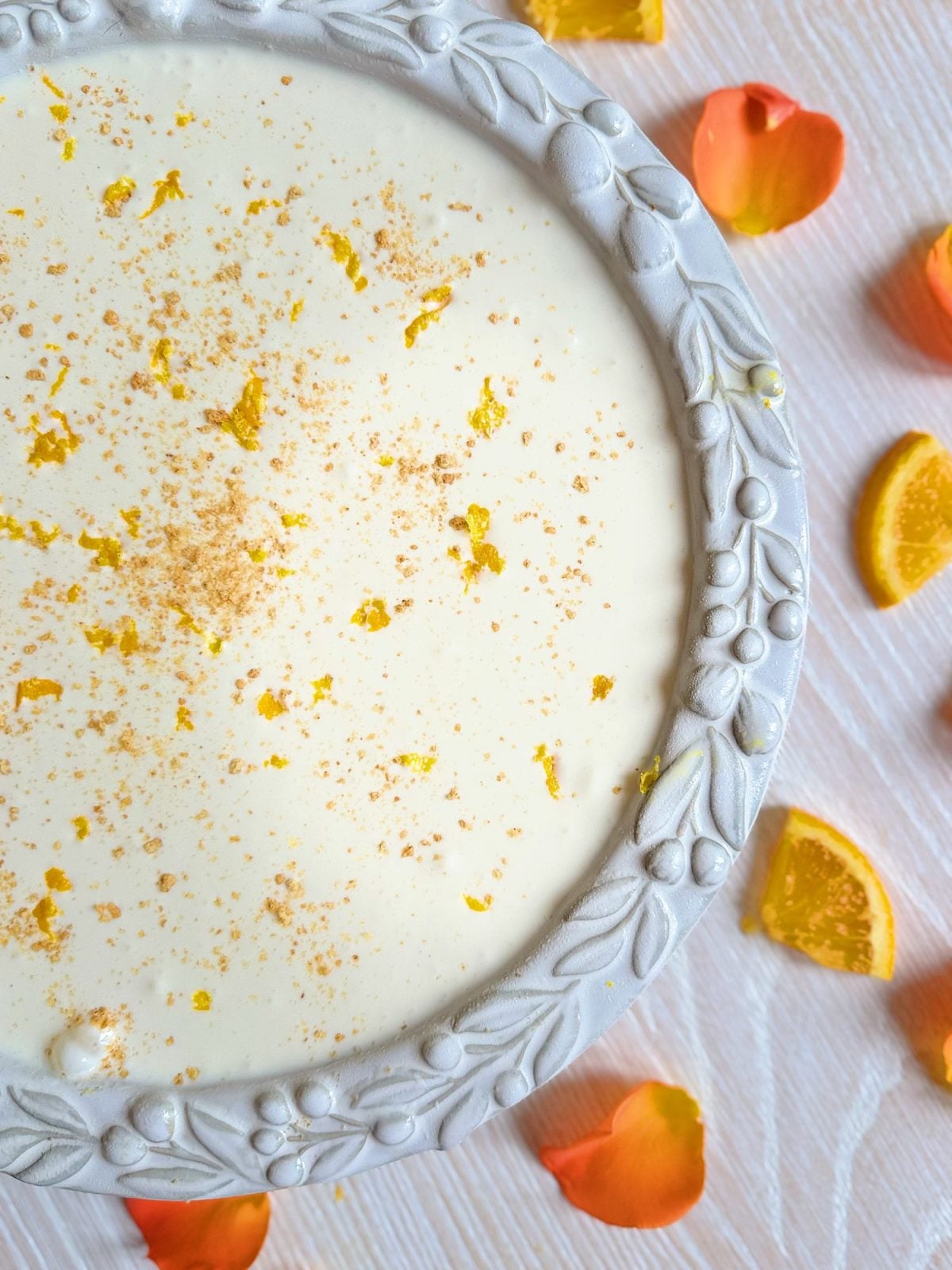 A white ceramic plate with a creamy dessert garnished with orange zest and a sprinkle of spices, surrounded by orange petals and slices on a light wooden surface.