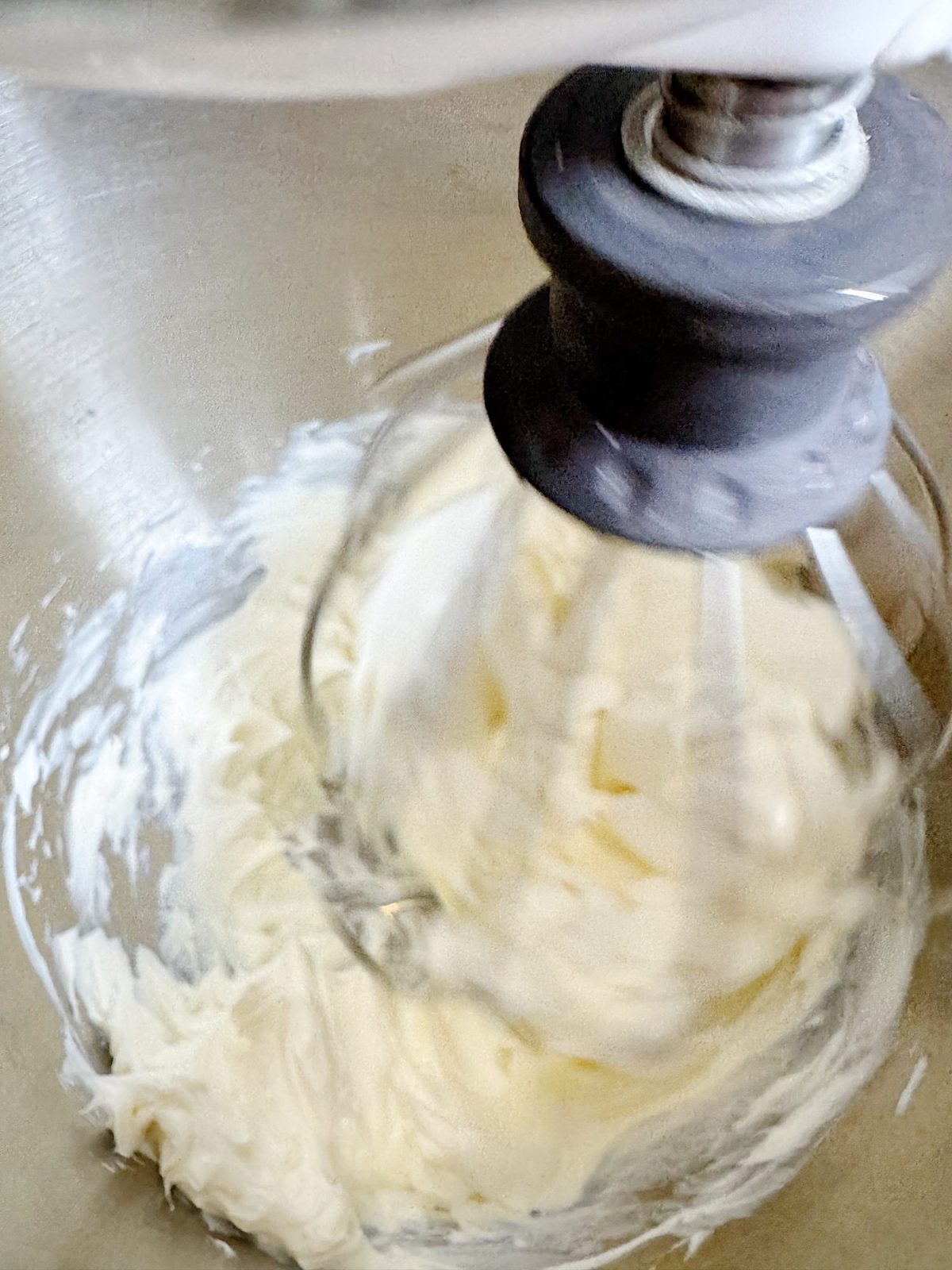 A stand mixer with a wire whisk attachment is whipping a creamy white mixture in a metal bowl.
