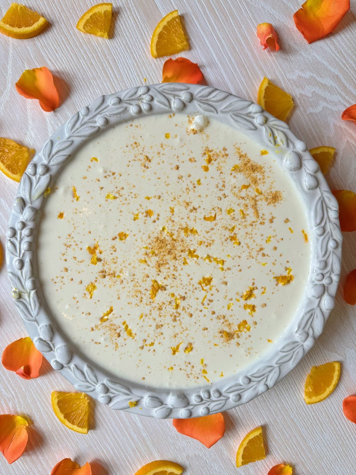 A round pie with a creamy white filling, garnished with orange zest, in a decorative dish. Orange slices and rose petals are scattered on a light wooden surface around the pie.