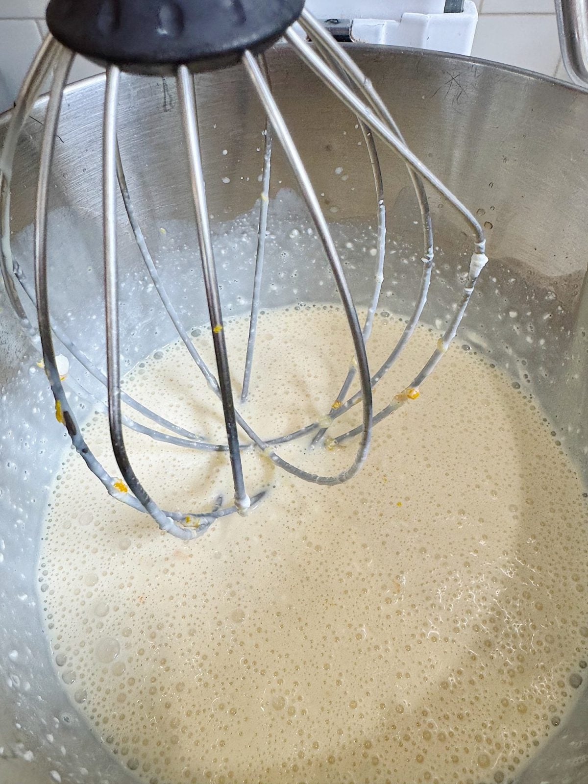A stand mixer with a whisk attachment mixing a pale, frothy batter in a stainless steel bowl.