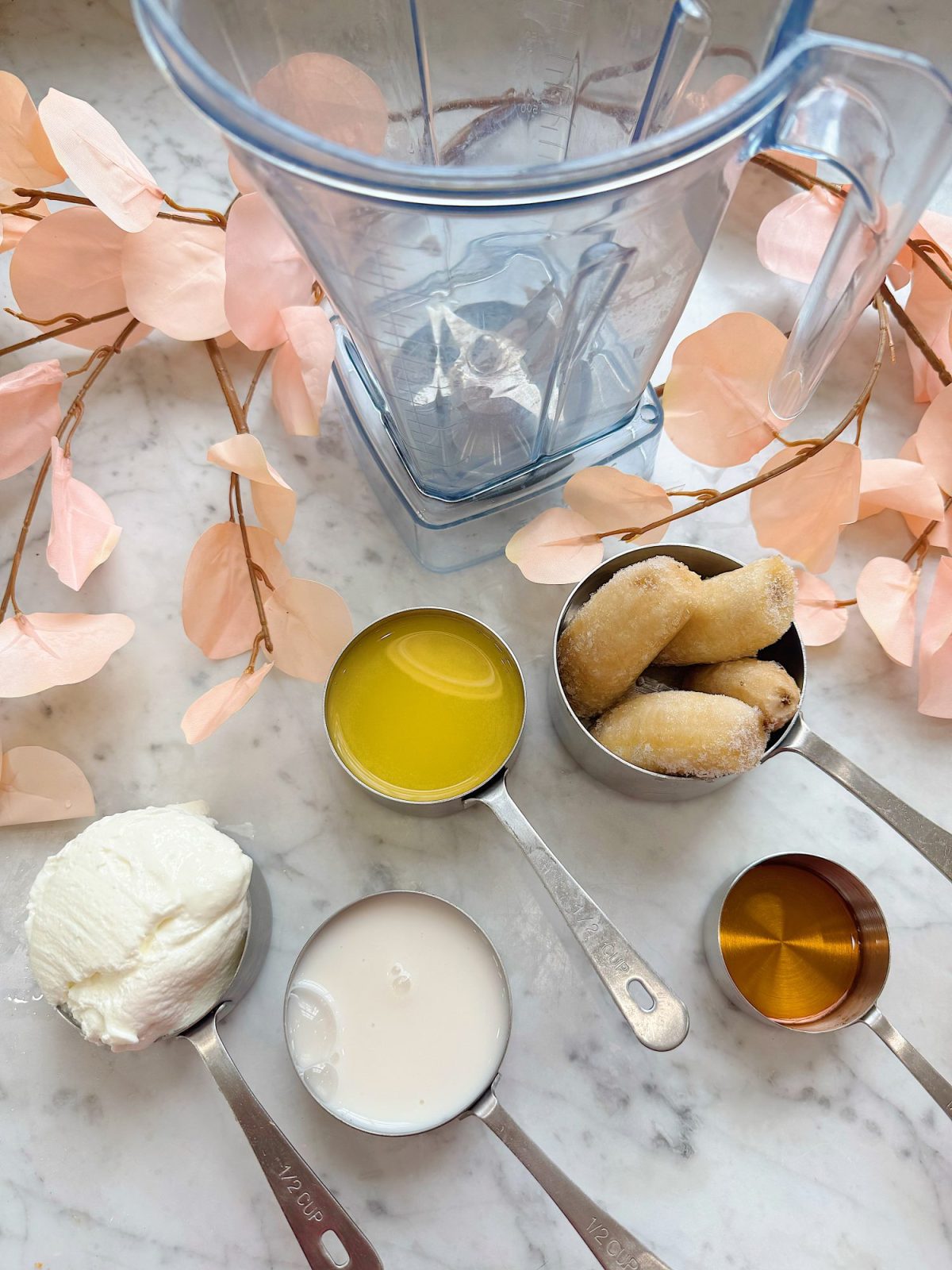 A blender with measuring cups containing frozen banana, melted butter, yogurt, milk, and syrup on a marble surface with pink decorative leaves.