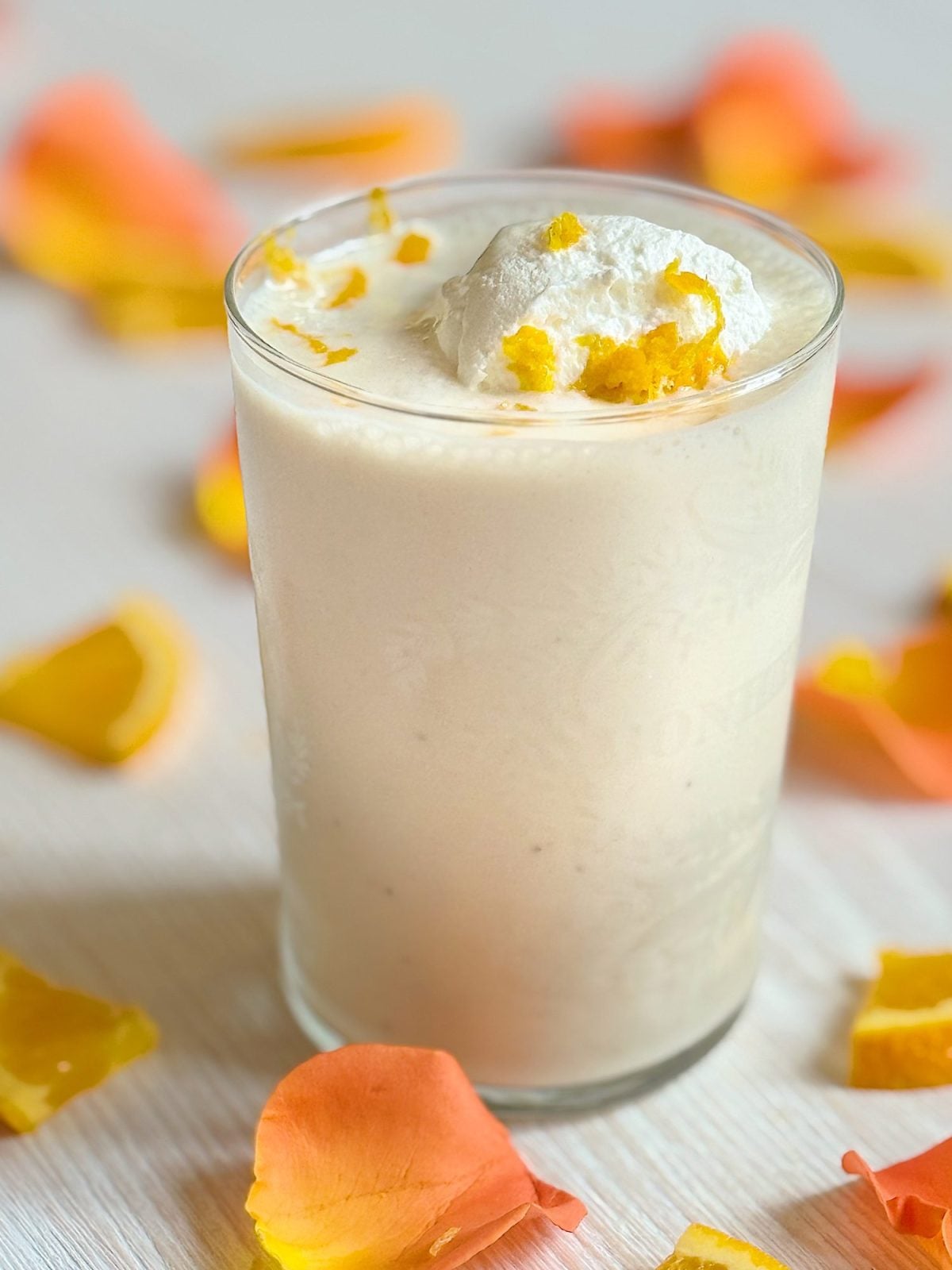 A glass of creamy beverage topped with whipped cream and orange zest, surrounded by scattered orange and peach-colored flower petals.