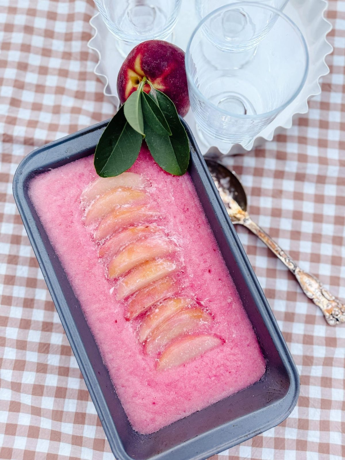 A rectangular dish of pink sorbet topped with sliced nectarines is placed on a checkered tablecloth, with a whole nectarine and empty glasses nearby.