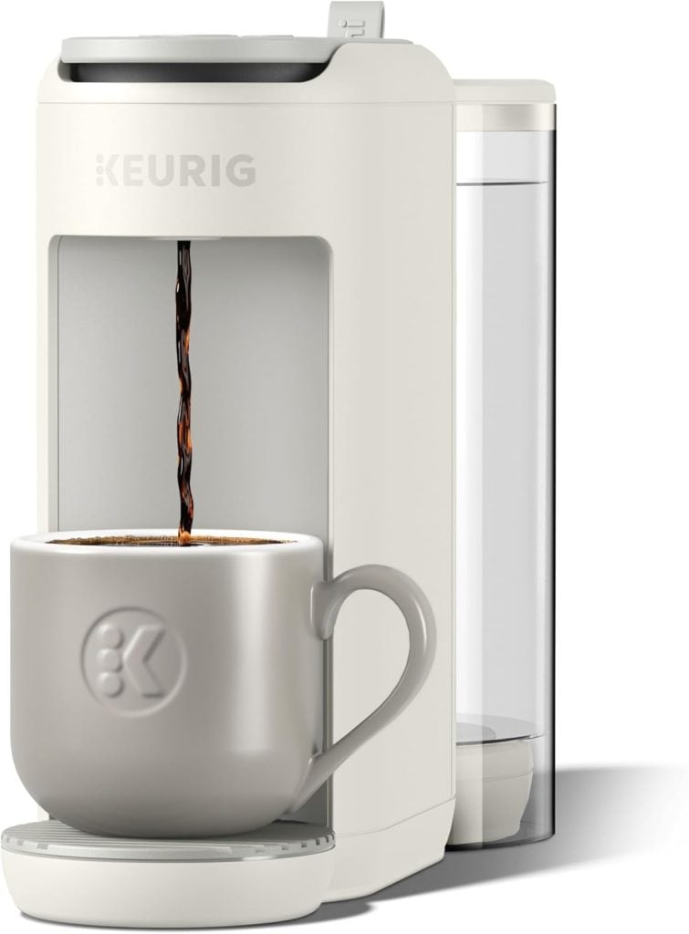 A white Keurig coffee maker brews coffee into a light gray mug with a logo, its clear water reservoir visible—perfect for upgrading your kitchen on Amazon Prime Day.