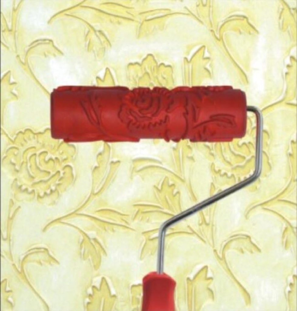A red patterned paint roller is held against a wall, creating a floral embossed design on a light yellow background—perfect for adding a touch of Creative Crafts to any space.