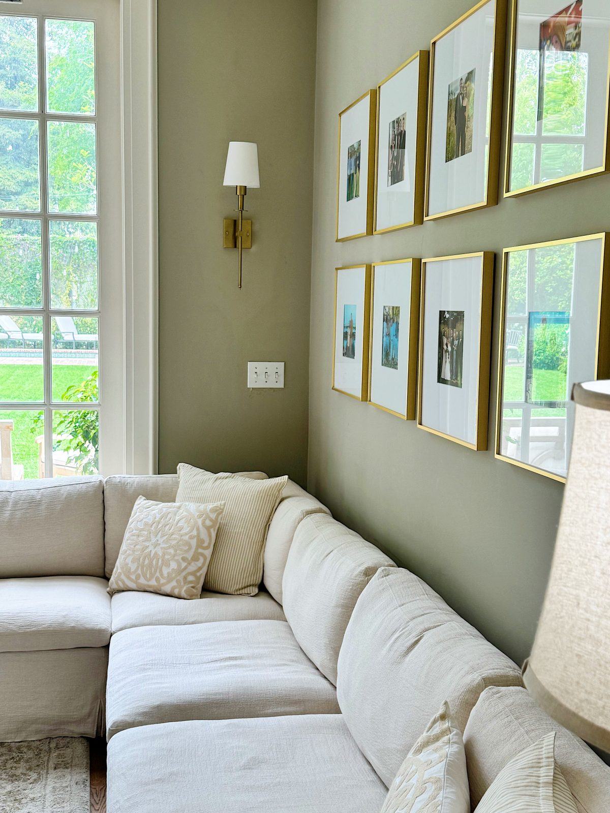 A beige sectional sofa with patterned pillows sits in a corner beneath framed photos and a wall sconce, next to large windows overlooking a green yard.