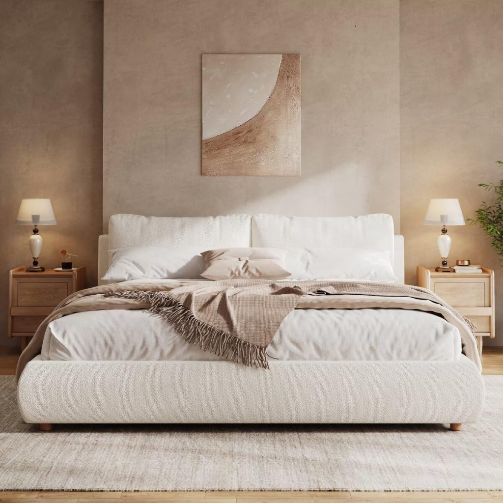 A modern bedroom with a large white bed, perfect for a restful summer sleep, light wood nightstands, matching lamps, a neutral abstract painting above the bed, and beige walls and rug.