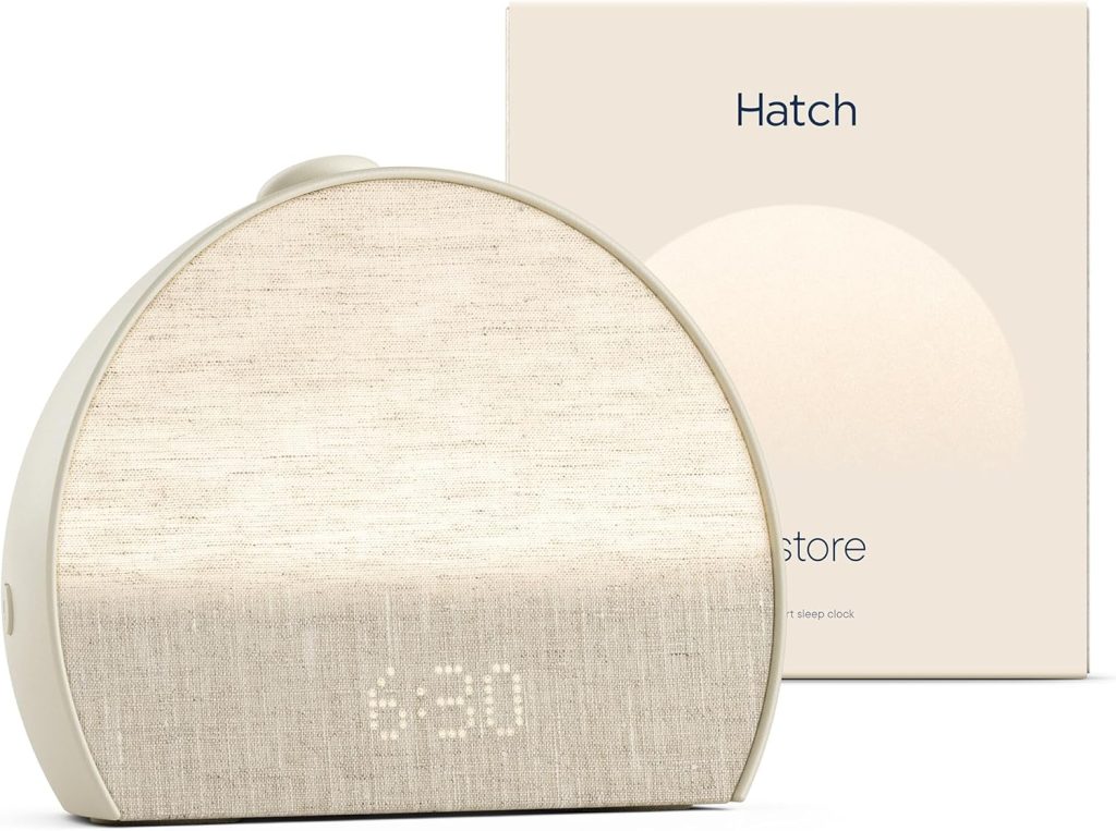 A Hatch Restore sunrise alarm clock with a soft fabric front and digital time display, perfect for refreshing summer sleep, positioned in front of its beige product packaging.