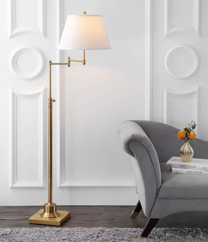 A gold floor lamp with a white shade stands next to a gray upholstered chair and a side table with books and a vase of orange flowers—perfect inspiration for your Amazon Prime Day home refresh, set against a white paneled wall.