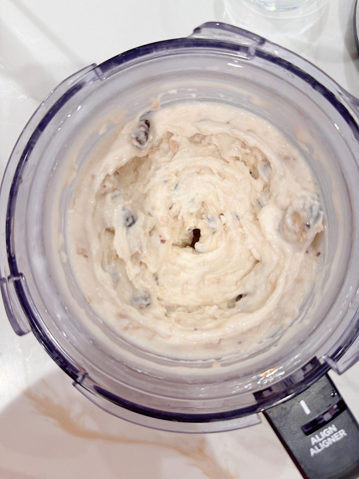 A food processor bowl containing a creamy, pale mixture with visible chunks, likely blended with add-ins such as nuts or chocolate chips.