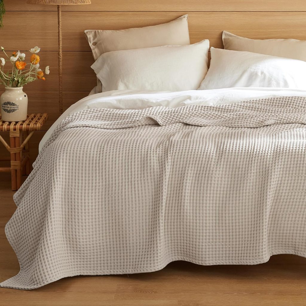 A neatly made bed with cream-colored pillows and a textured beige blanket promises restful summer sleep, set against a wooden headboard with a small table and a vase of flowers beside it.