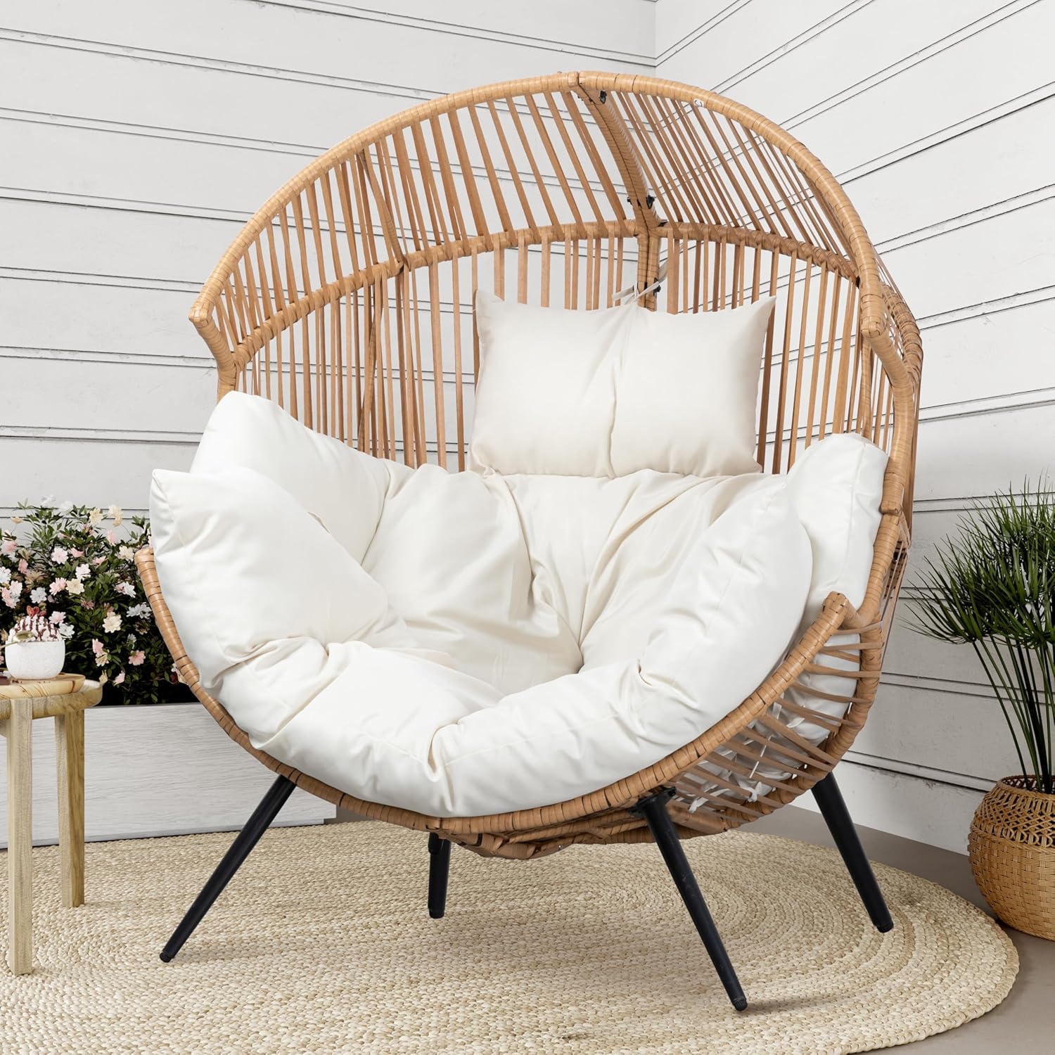 A round rattan chair with white cushions and a pillow sits on a woven rug in a room with white paneled walls, a potted plant, and a small side table with Amazon Prime flowers.