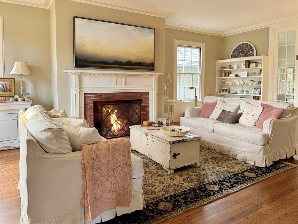 A cozy living room with a lit fireplace, two white sofas, a rustic coffee table, built-in bookshelves, and framed artwork above the mantle.
