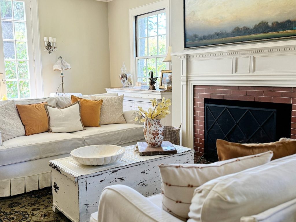A bright living room with white sofas, brown and white pillows, a rustic coffee table, fireplace, large painting, and a window letting in natural light.