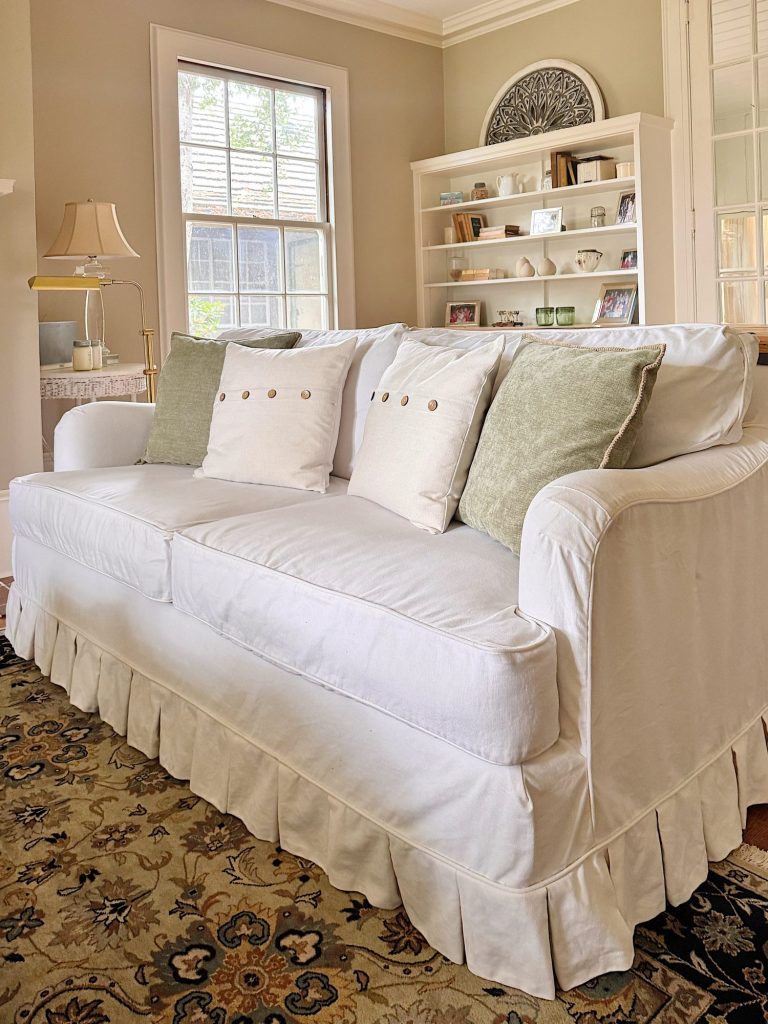 A white sofa with three cushions and two green pillows sits in a living room with a patterned rug, bookshelves, a lamp, and large windows in the background.