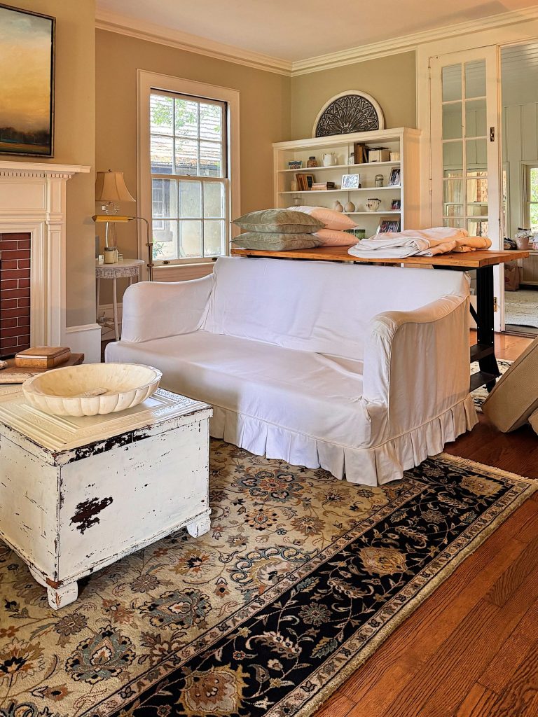 A living room with a white slipcovered sofa, a distressed white coffee table, a patterned rug, and bookshelves against beige walls with a window and French doors in the background.