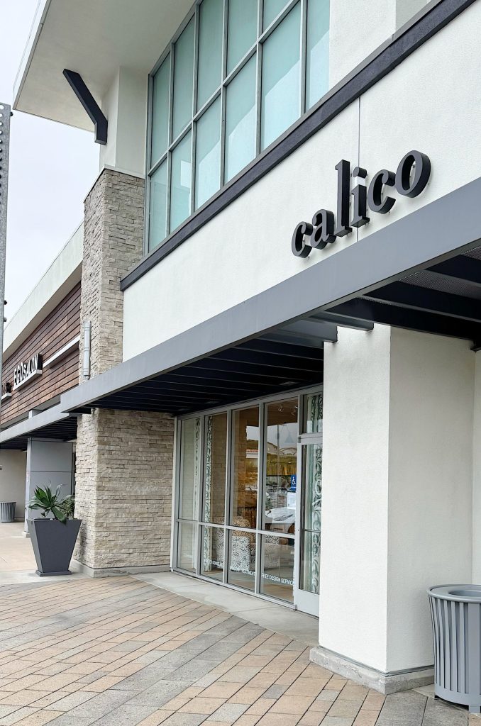 Storefront of Calico with large windows, a visible doorway, modern exterior design, and a sidewalk with potted plants outside.