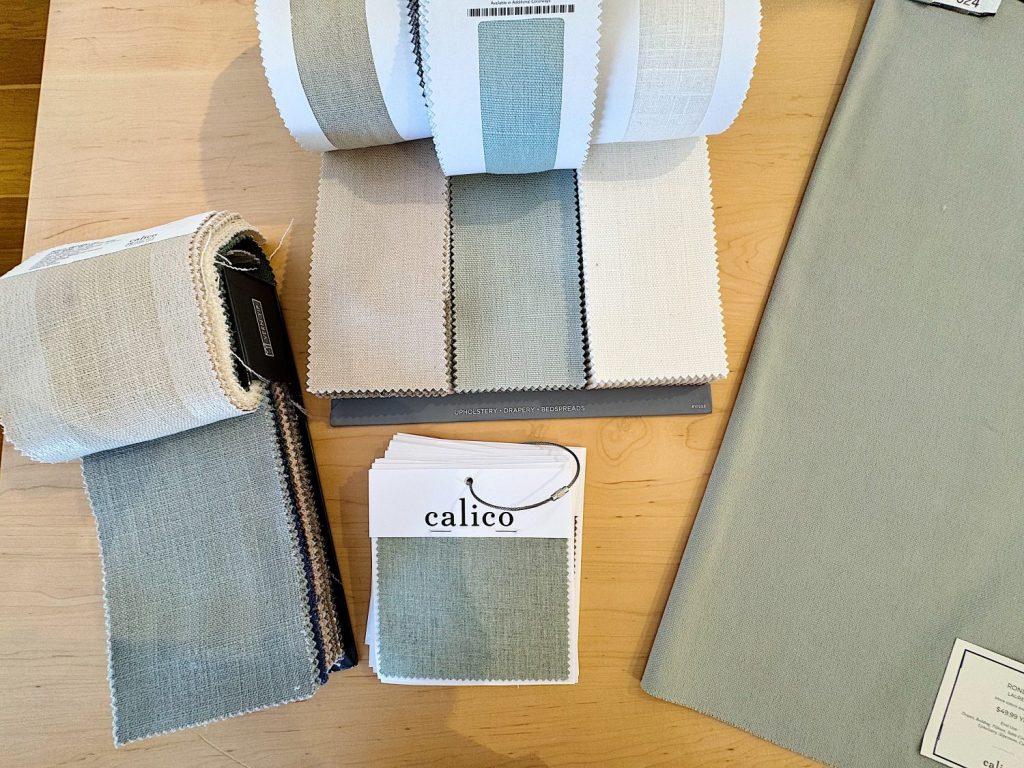 Fabric swatches and sample books in neutral and light blue tones are arranged on a wooden table, including one labeled "calico.