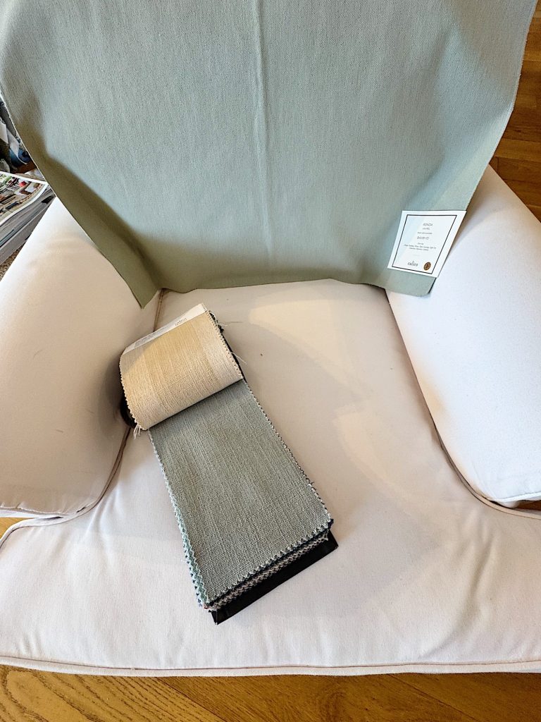 A white upholstered chair with a fabric sample booklet in neutral shades placed on the seat and a light green fabric draped over the backrest.