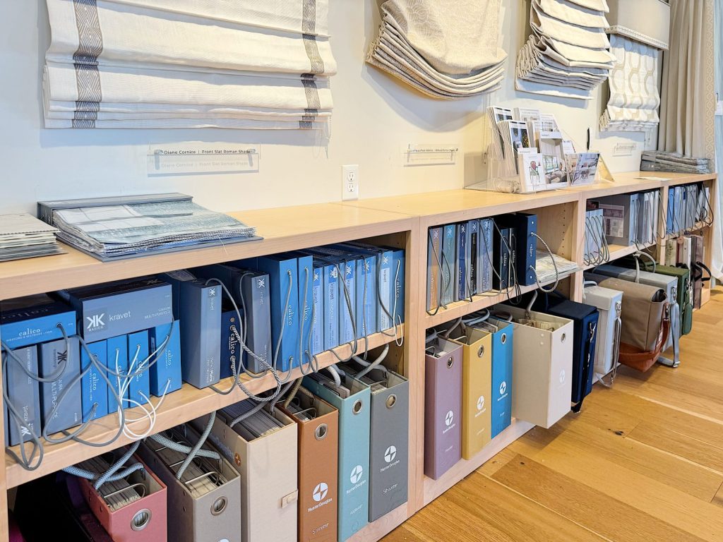 Shelves with organized fabric sample books and binders, brochures on the countertop, and Roman shades displayed above on the wall.