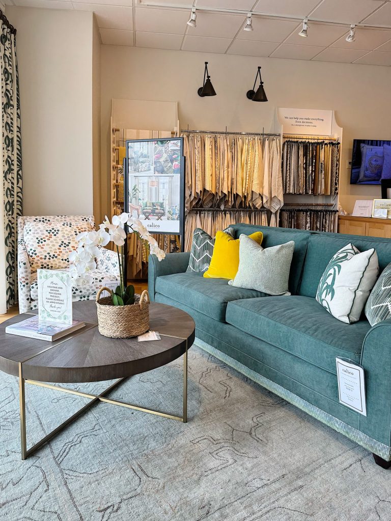 A showroom features a teal sofa with yellow and patterned pillows, a round coffee table with decor, a patterned armchair, and fabric samples displayed in the background.