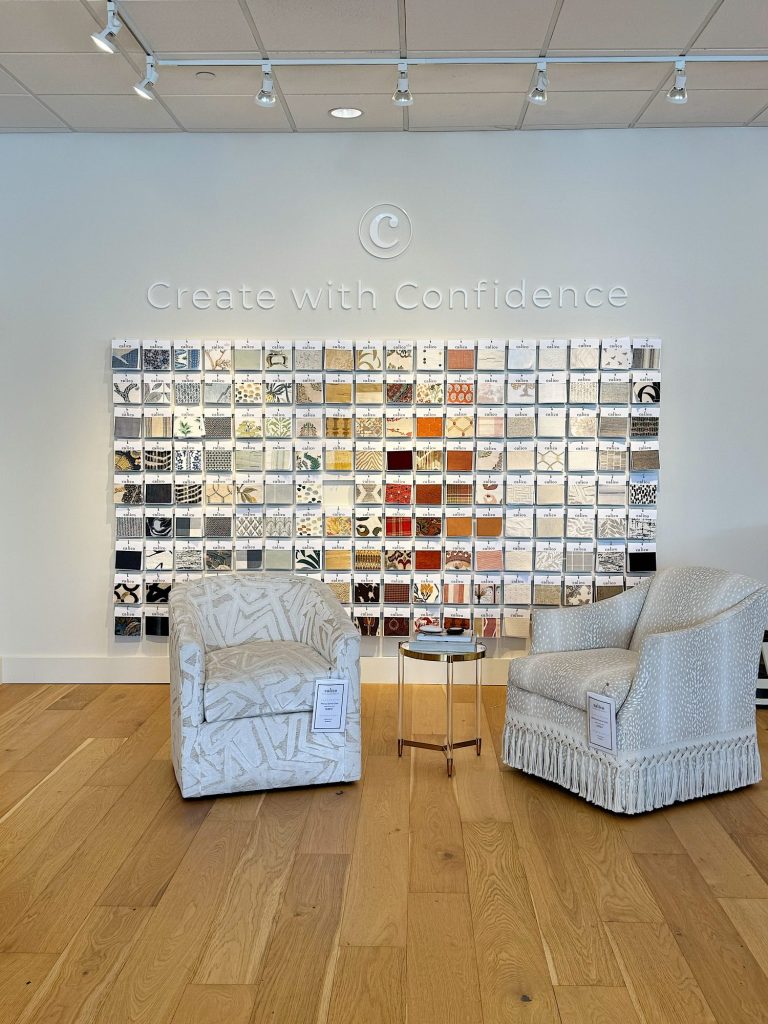 Two upholstered chairs and a small round table are arranged on a wooden floor in front of a wall display of fabric samples with the words "Create with Confidence" above.
