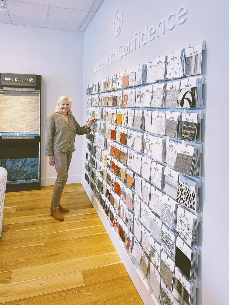 A woman stands by a display wall of fabric samples in a showroom, touching one sample. The wall has the phrase "Create with confidence" above the samples.