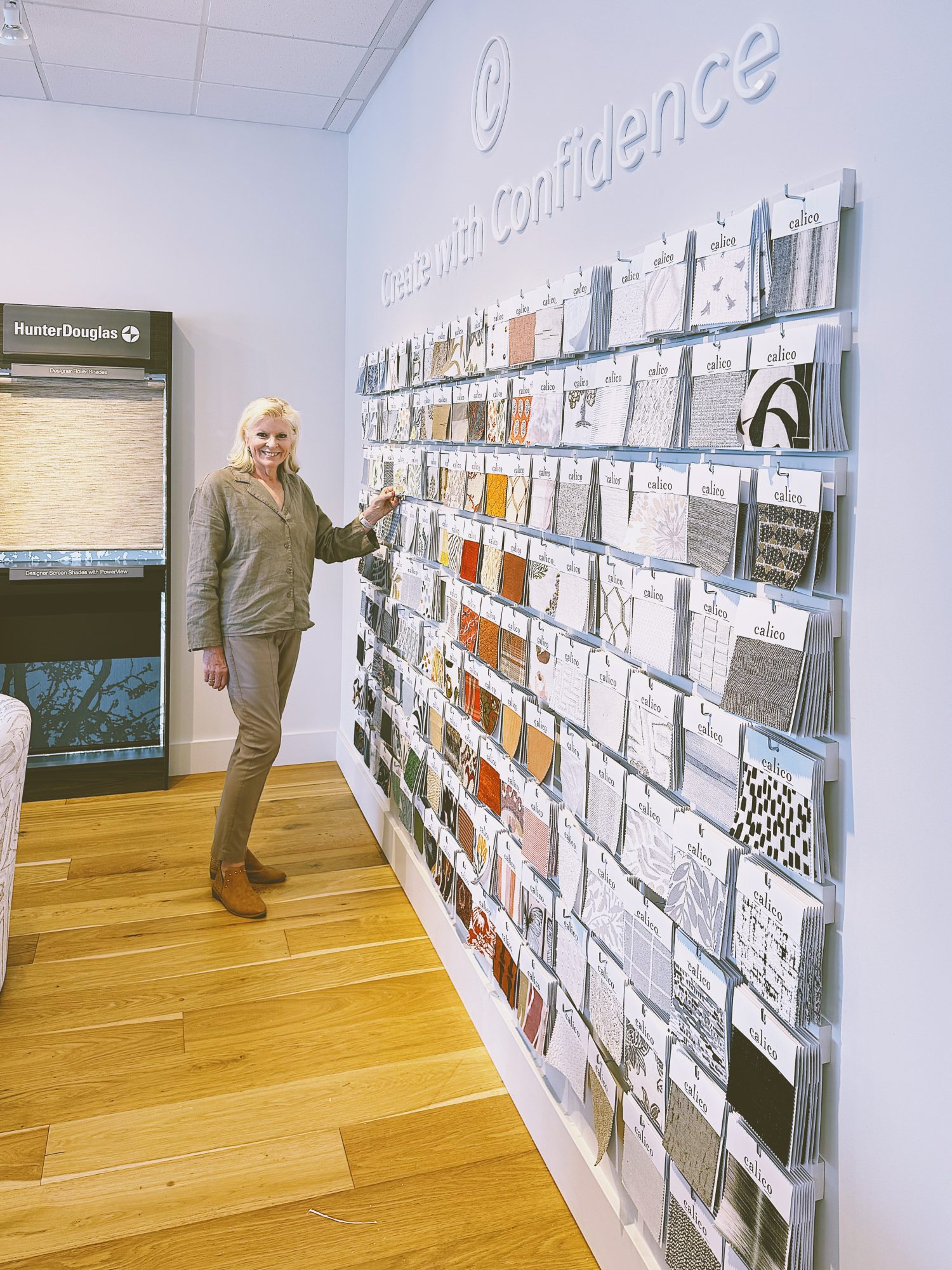 A woman stands by a display wall of fabric samples in a showroom, touching one sample. The wall has the phrase "Create with confidence" above the samples.