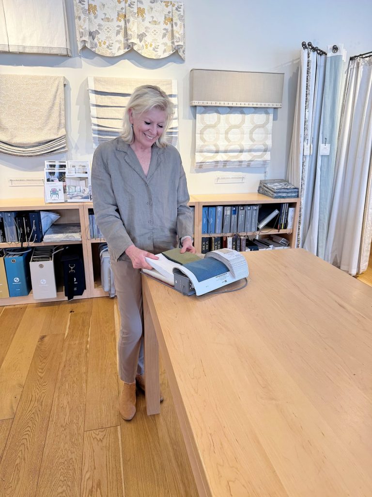 A woman stands at a wooden table, operating a fabric-cutting machine in a room with fabric samples and bookshelves in the background.