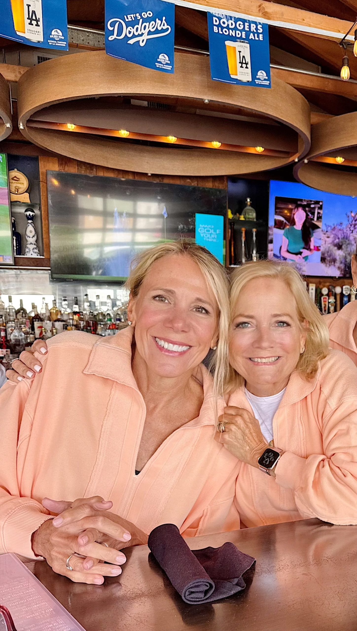 Two women wearing matching peach-colored tops sit closely together at a bar, smiling at the camera. Dodgers signs and drinks are visible in the background.