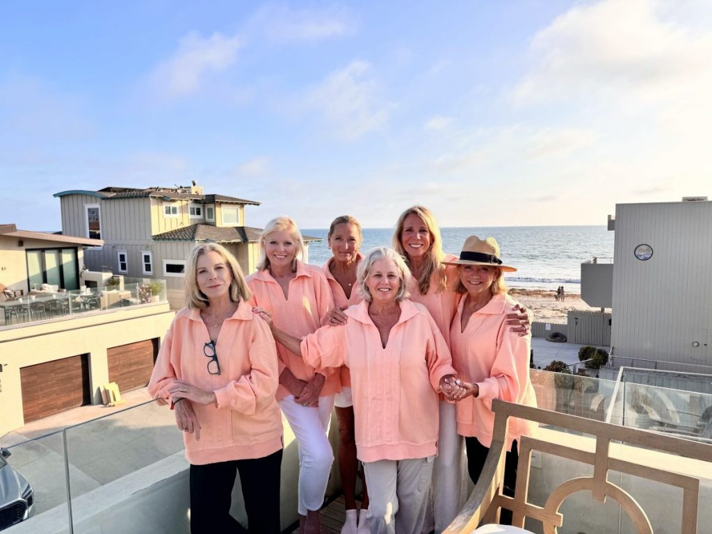 Six women wearing matching peach-colored tops stand together on a balcony overlooking the beach, smiling at the camera with houses and the ocean in the background.