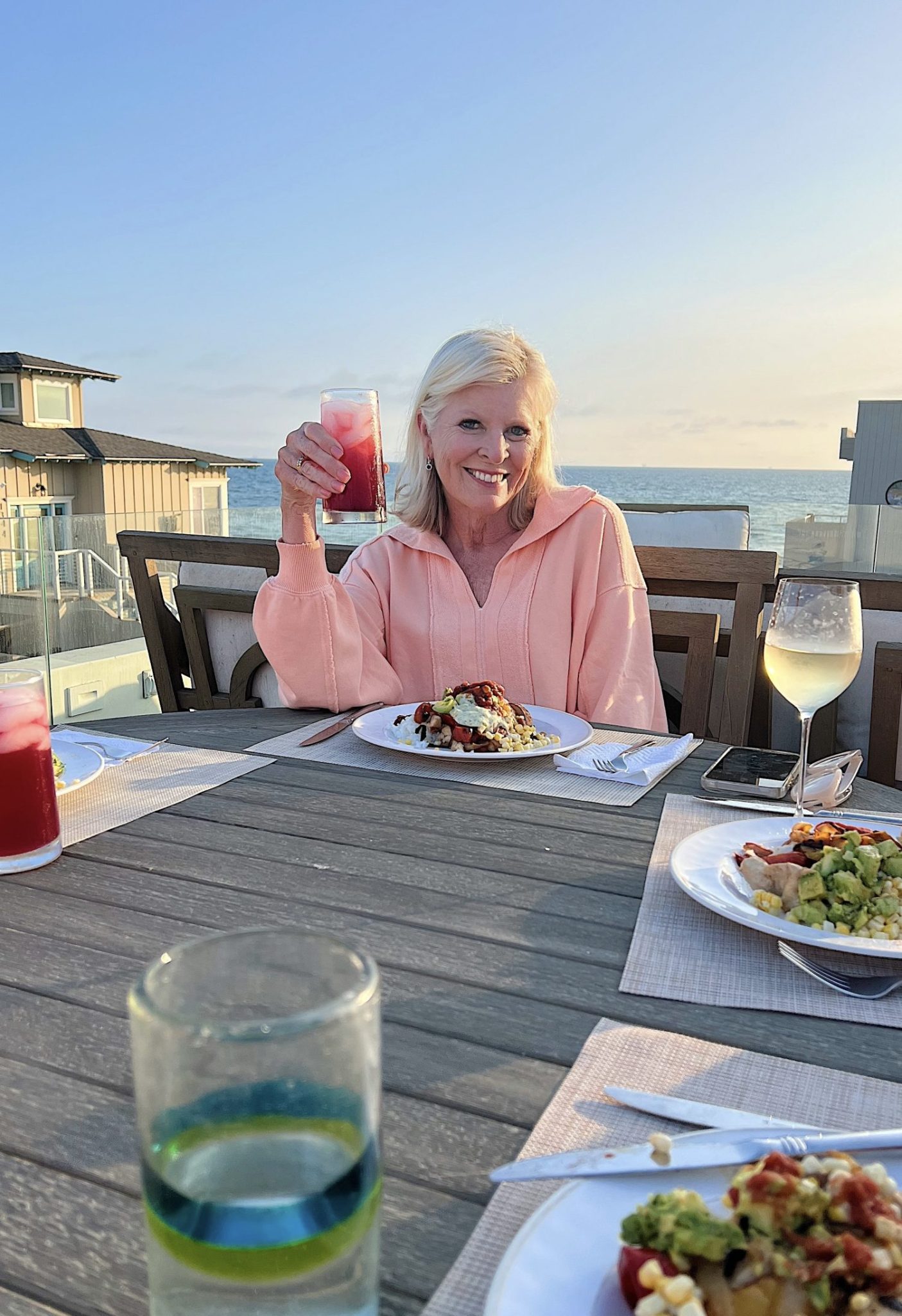 Older woman with white hair raises a red drink while seated at an outdoor table set for a meal by the ocean, with food and drinks visible on the table.